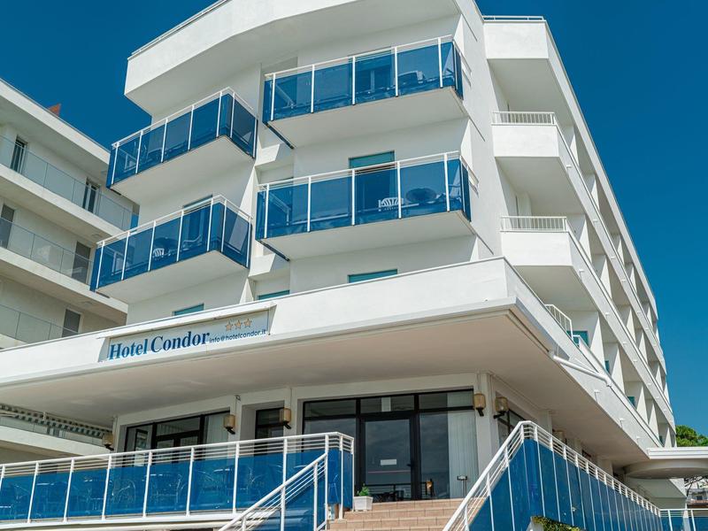 Edificio d'hotel bianco moderno con balconi in vetro sotto un cielo azzurro.