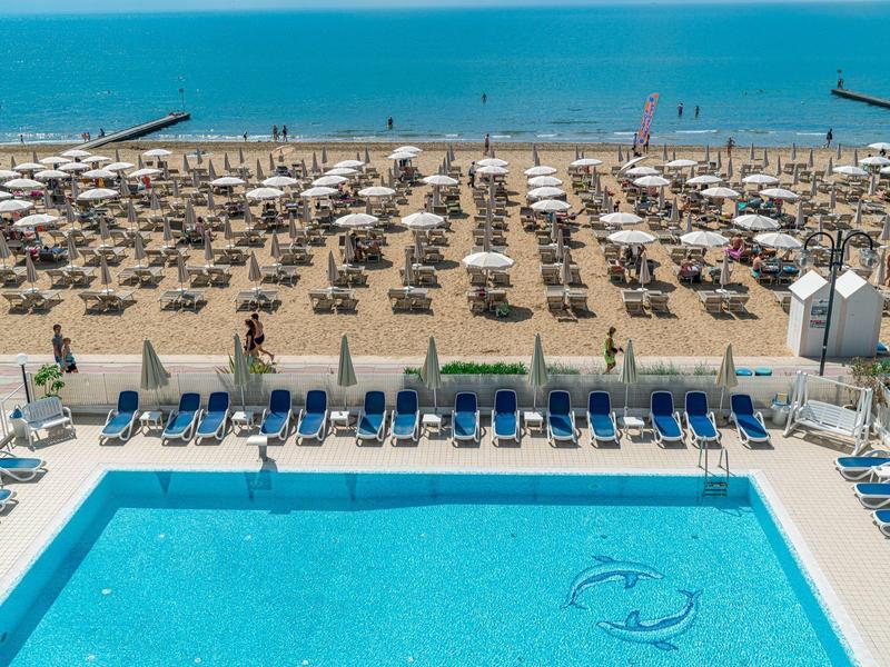 Vista di una piscina con lettini, dietro una spiaggia di sabbia con ombrelloni e il mare sullo sfondo.