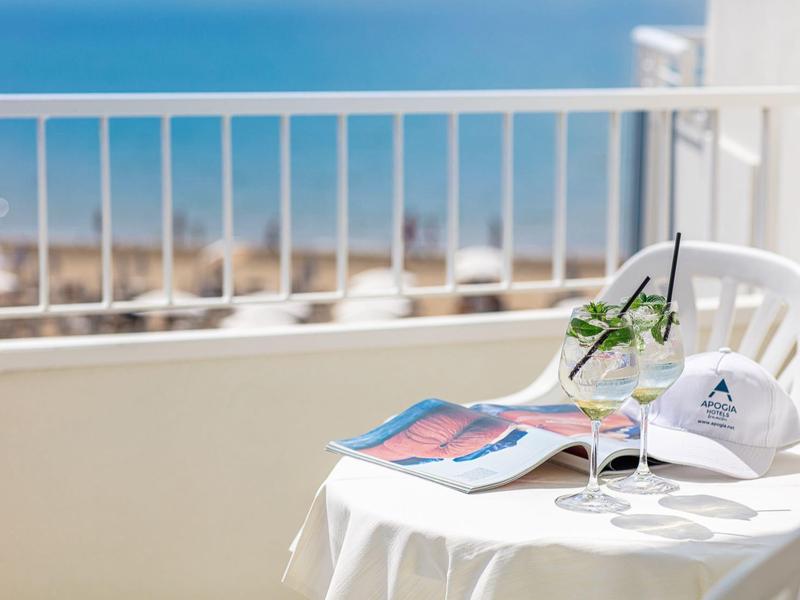 Tavolo bianco con bevanda, rivista e cappello su balcone con vista mare.