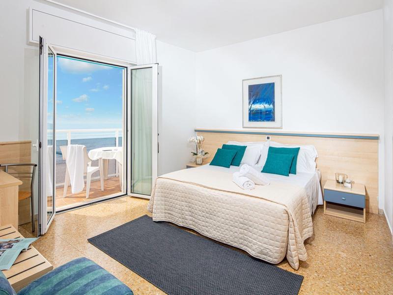 Camera d'hotel luminosa con letto matrimoniale, accenti blu e balcone con vista sul mare.