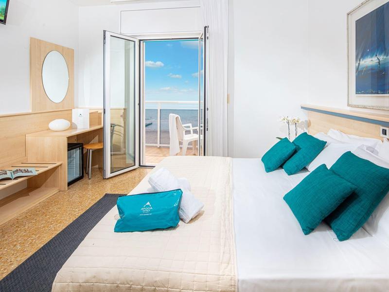 Camera d'hotel moderna con letto matrimoniale, balcone e vista mare sotto un cielo limpido.