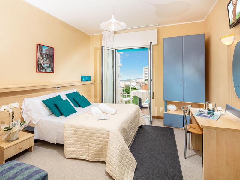 Camera d'hotel luminosa con letto matrimoniale, armadio blu, scrivania e balcone con vista sulla città.