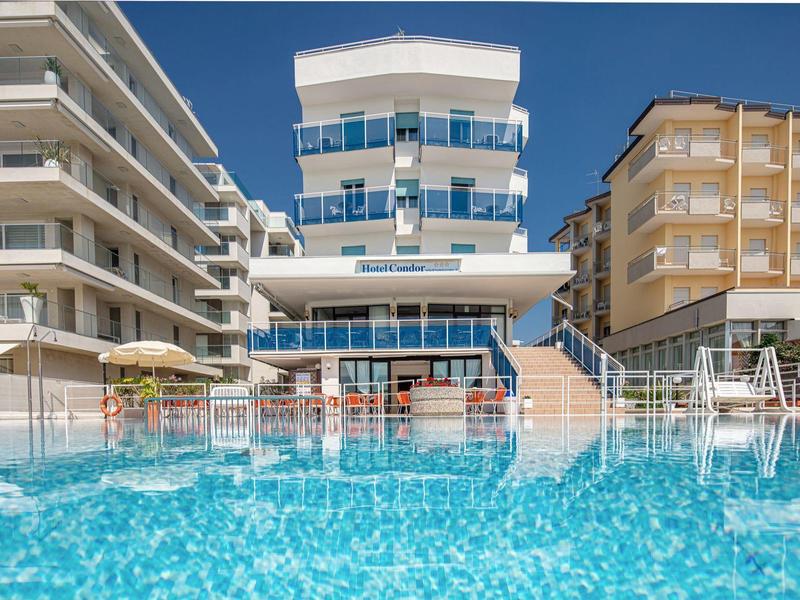 Hotel moderno con piscina e ombrelloni, circondato da altri edifici sotto un cielo sereno.