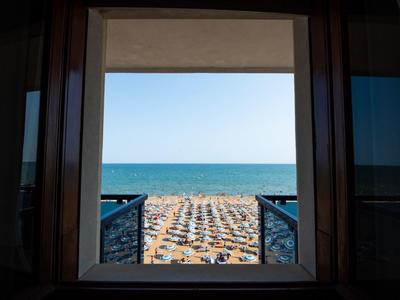 Widok przez okno na zatłoczoną plażę z parasolami i morzem w tle.