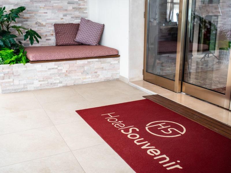 Wejście z czerwonym dywanikiem i ławką z poduszkami obok szklanych drzwi.