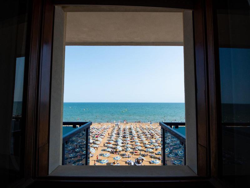 Widok przez okno na zatłoczoną plażę z parasolami i morzem w tle.
