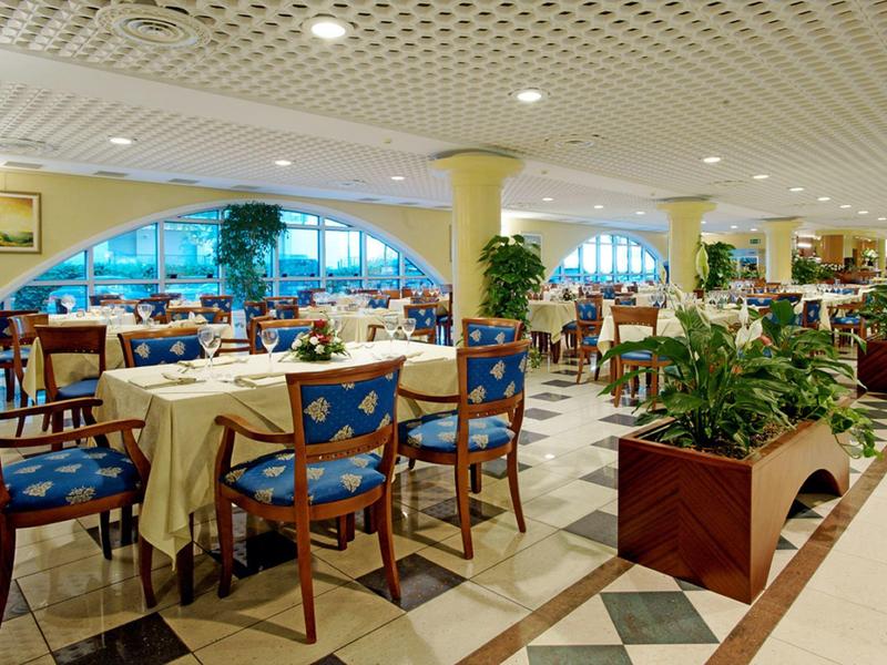 Grote hotel eetzaal met blauwe stoelen, witte tafelkleden en veel planten.