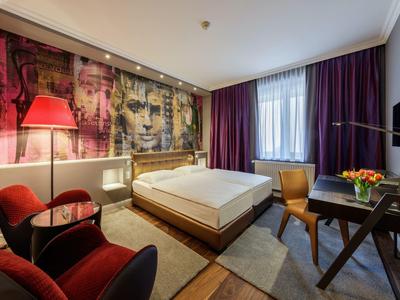 Modern eingerichtetes Hotelzimmer mit Doppelbett, buntem Wandbild, roten Sesseln und Fenster mit Vorhängen.