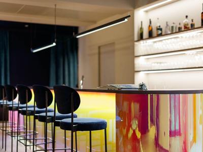Moderner Hotelbarbereich mit leuchtender Theke und hohen Barstühlen vor dunklen Vorhängen.