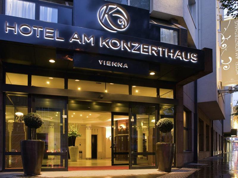 Eingang eines Hotels mit hellem Beleuchtung, großen Glas- und Metallelementen, zwei Pflanzenkübeln.