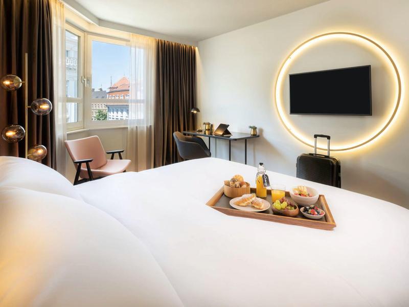 Hell eingerichtetes Hotelzimmer mit großem Bett, bestrahltem runden Spiegel und Fenster mit braunen Vorhängen.