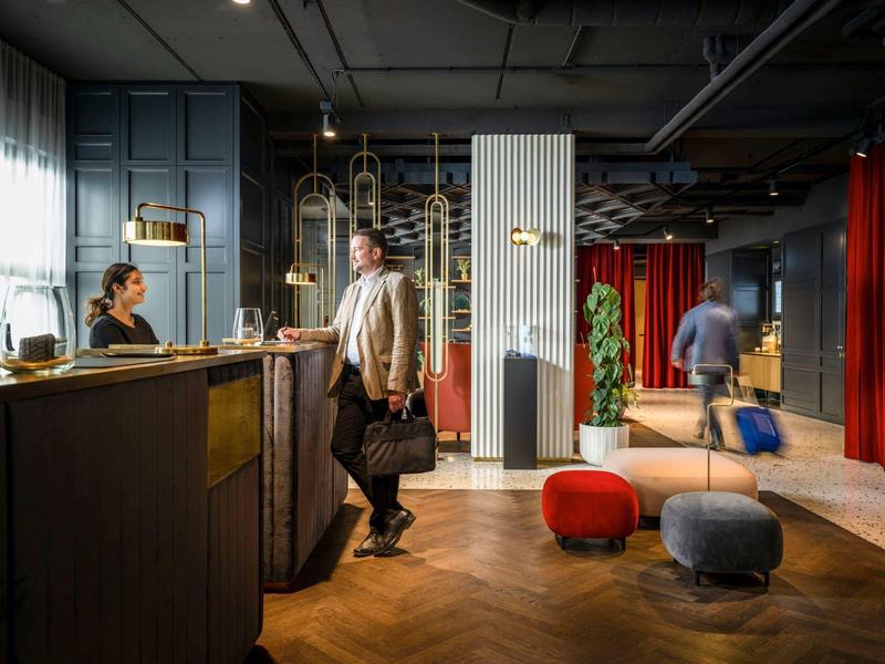 Moderne Hotelrezeption mit Gästen, dunklem Holz, Teppichboden und gemütlichen Sitzgelegenheiten.