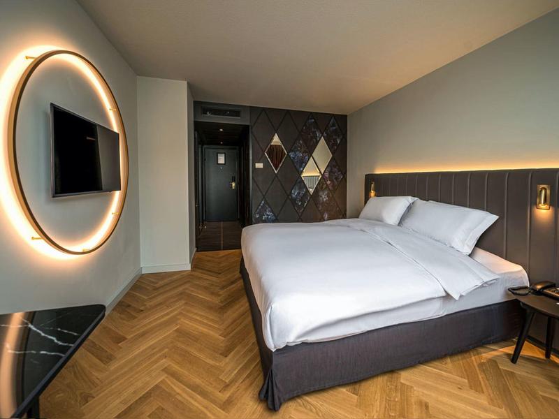 Modernes Hotelzimmer mit Doppelbett, Holzboden, großem runden Spiegel und LED-Beleuchtung.