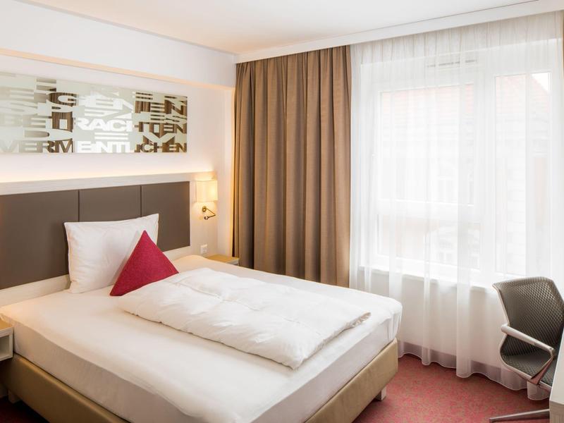 Gemütliches Einzelzimmer mit Bett, rotem Kissen, Schreibtisch und großem Fenster mit Vorhängen.