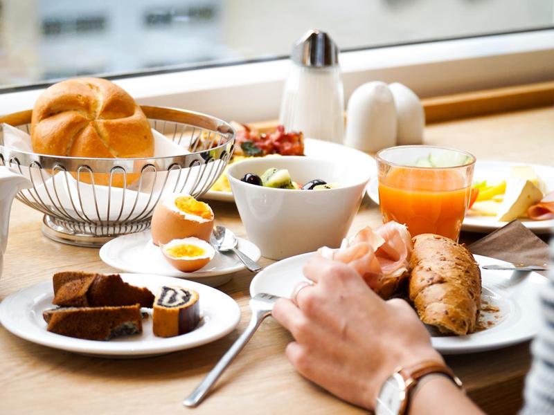 Frühstückstisch mit Brötchen, gekochtem Ei, Aufschnitt und Orangensaft vor einem Fenster.