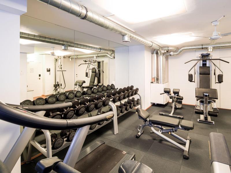 Kleines Fitnessstudio mit Hanteln, Trainingsbänken und verschiedenen Geräten in einem hellen Raum.