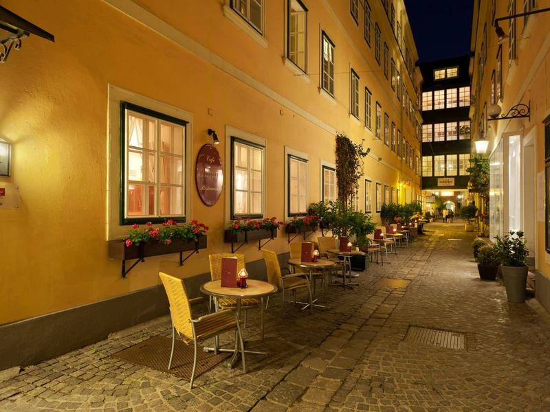 Enge Gasse mit gelben Häusern, beleuchteten Fenstern und kleinen Tischen mit Stühlen am Gehweg.