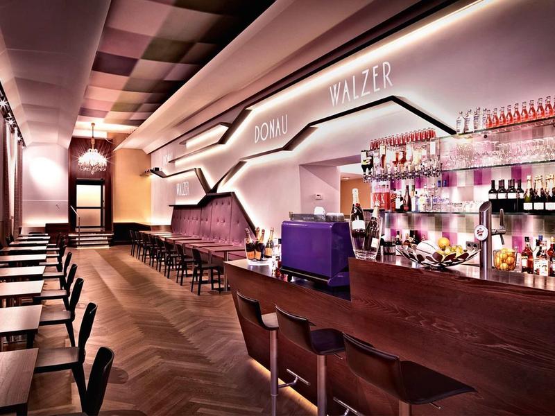 Moderner Barraum mit langer Holztheke, hohen Hockern, indirekter Beleuchtung und dunklem Holzboden.