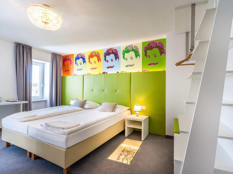 Helles Schlafzimmer mit zwei Einzelbetten, grünem Kopfteil, bunten Portraits und weißem Nachttisch.