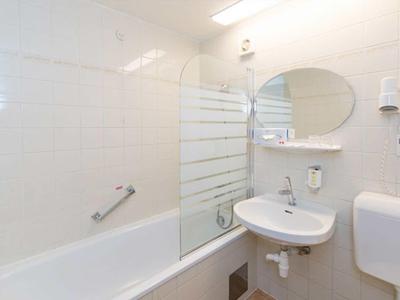 Helles Badezimmer mit Badewanne, Glas-Duschwand, rundem Spiegel, Waschbecken und Toilettenspülkasten.