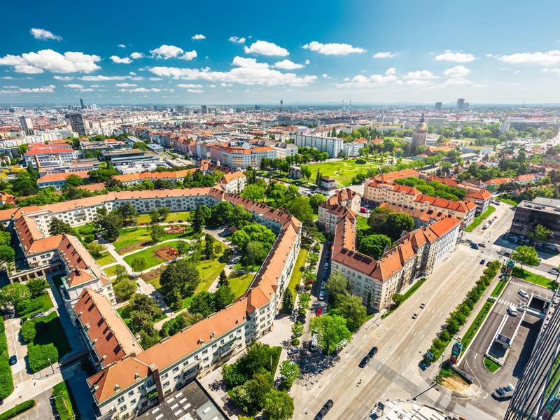 Luftaufnahme einer Stadt mit grünen Parks, roten Dächern und breiten Straßen unter blauem Himmel.
