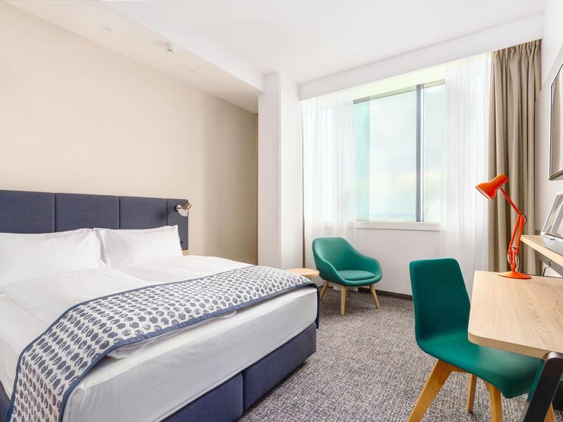 Helles Hotelzimmer mit Doppelbett, Schreibtisch, zwei grünen Stühlen und großem Fenster.