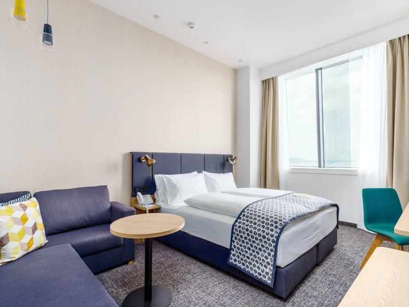 Modernes Hotelzimmer mit großem Bett, blauem Sofa, kleinem Tisch und großem Fenster mit Vorhängen.