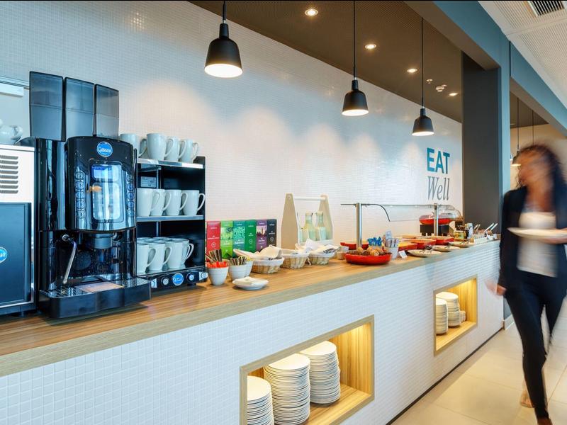 Moderne Hotelküche mit Kaffeeautomaten und Frühstückszutaten, Person läuft vorbei.