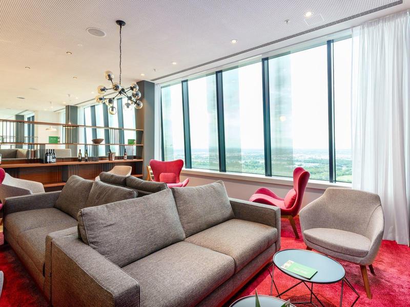 Modernes Hotelzimmer mit grauem Sofa, roten Sesseln und großen Panoramafenstern mit Ausblick.