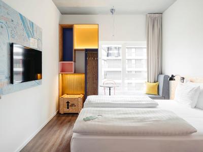 Helles Hotelzimmer mit Doppelbett, großem Fenster, TV an der Wand und modernem Holzregal.