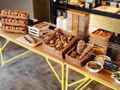 Tisch mit Holzboxen voller Bagels, Brote und Müsli im hellen Raum mit Betonboden.
