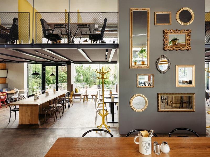 Moderner Gastraum mit Holztischen, Stühlen, großen Fenstern und dekorativen Spiegeln an der Wand.