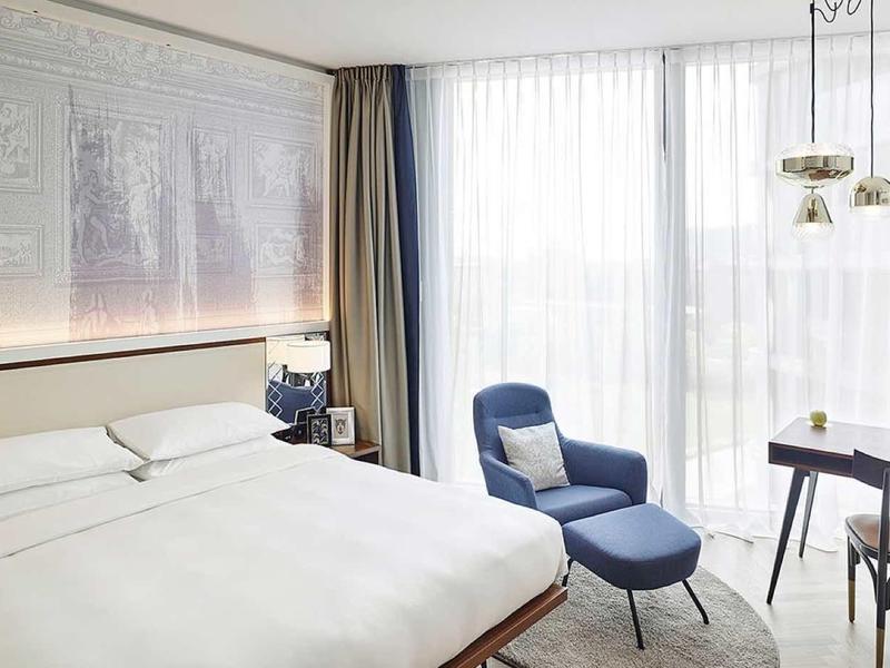 Jasny, nowoczesny pokój hotelowy z dużym łóżkiem, miejscem do siedzenia i biurkiem przy oknach od podłogi do sufitu.