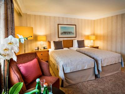 Světý hotelový pokoj se dvěma jednolůžky, nočními stolky, lampami a posezením s červenými polštáři.