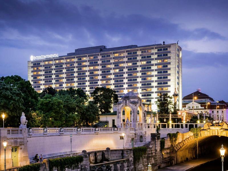 Velká hotelová budova osvětlená za soumraku u řeky s kamenným mostem před ní.