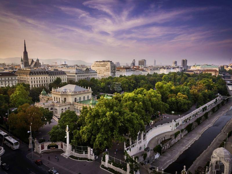 Městský park s stromy, řekou a historickými budovami pod fialovou oblačnou oblohou za soumraku.