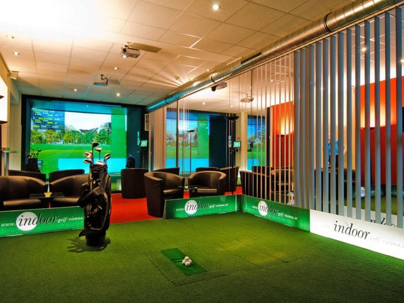 Nowoczesny kryty teren golfowy z sztuczną trawą, fotelami i podświetlanymi reklamami.