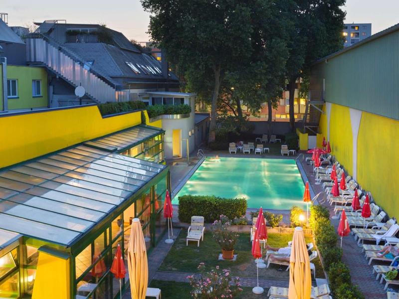 Oświetlony basen z leżakami i parasolami na dziedzińcu hotelu o zmierzchu.