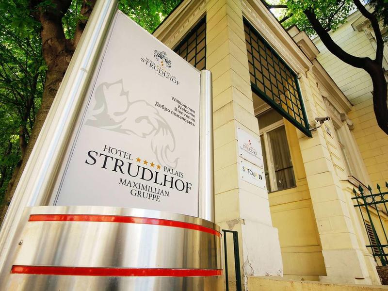 Eingang mit weißem Schild und rotem Band vor historischem gelbem Gebäudefassaden mit Bäumen.