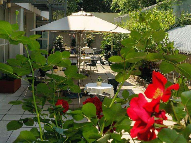 Garten mit rotem Blumenbusch, gepflastertem Weg und Sitzbereich unter weißem Sonnenschirm.