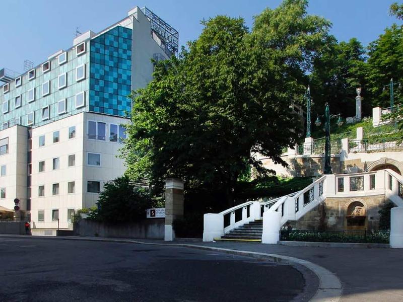 Städtische Straße mit mehrstöckigem Gebäude links und heller Treppe mit Grün rechts im Sonnenlicht.