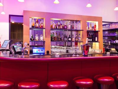 Nowoczesny bar hotelowy z czerwonymi stołkami, podświetlanym barem i półkami pełnymi butelek.