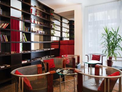 Przytulny salon hotelowy z czerwonymi fotelami, szklanymi stolikami i dużą półką na książki.