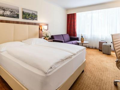 Licht hotelkamer met groot bed, bank, bureau en raam met gordijnen.