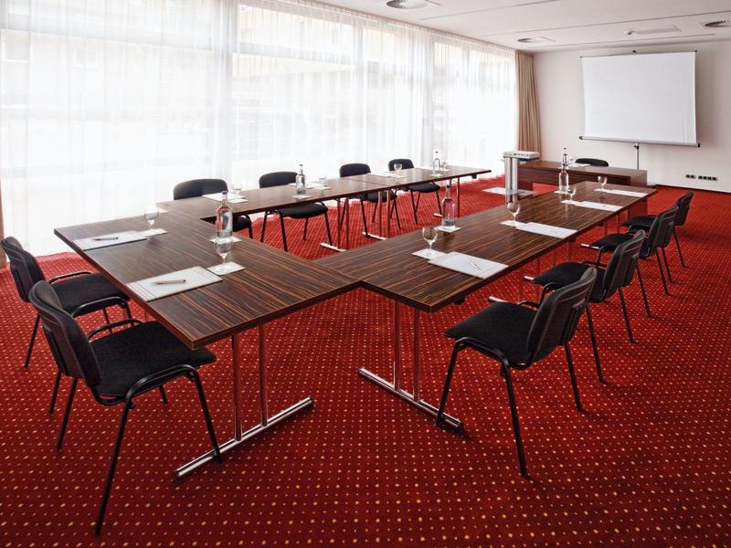 Konferenční místnost s U-tvarovaným stolem, černými židlemi a červeným kobercem.