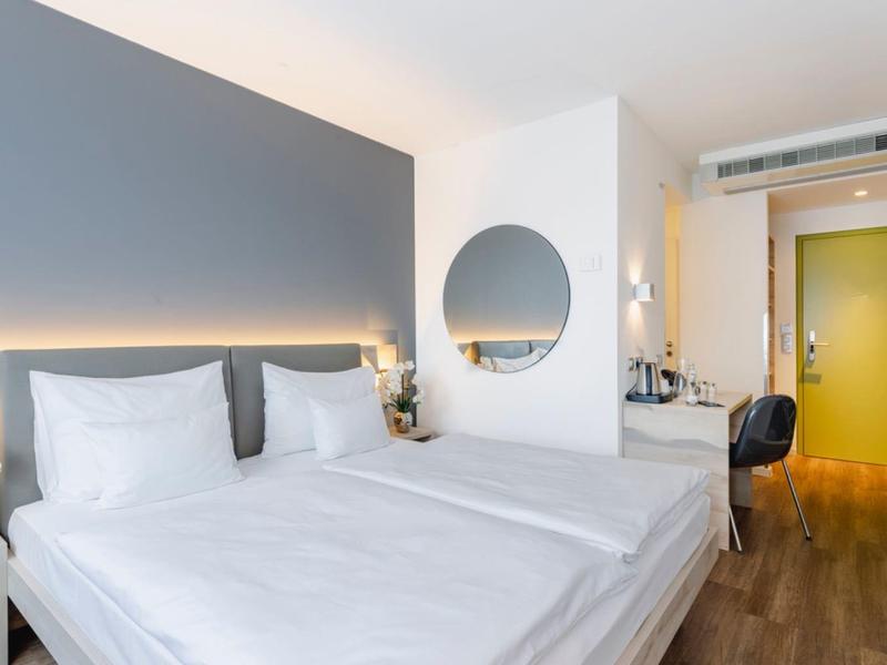 Moderní hotelový pokoj s manželskou postelí, kulatým zrcadlem a dřevěnou podlahou.