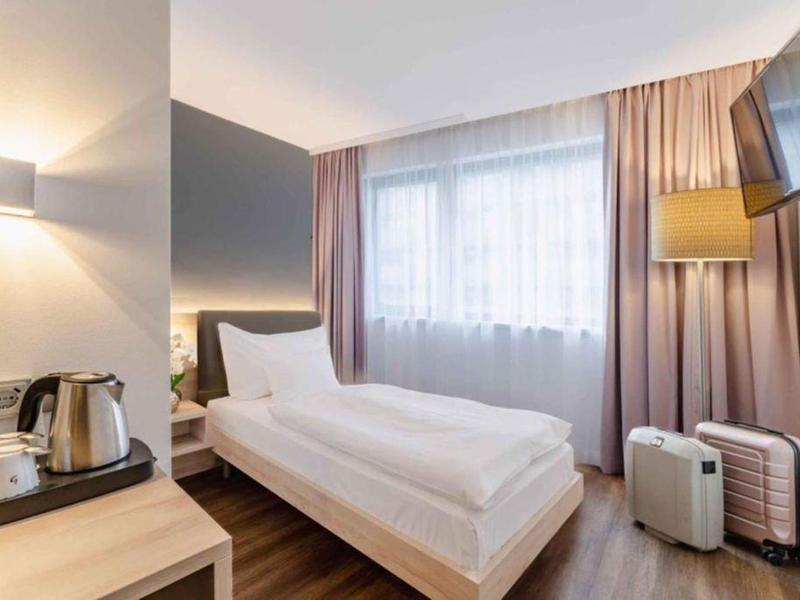 Světý hotelový pokoj s jednolůžkem, oknem, závěsy a stojícím kufrem.