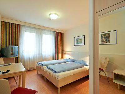 Hell eingerichtetes Hotelzimmer mit Doppelbett, Tisch, Stuhl, Fernseher und großen Fenstern mit Vorhängen.