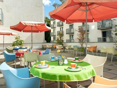 Kolorowy taras na zewnątrz z stołami, krzesłami i parasolami na tle nowoczesnych budynków.