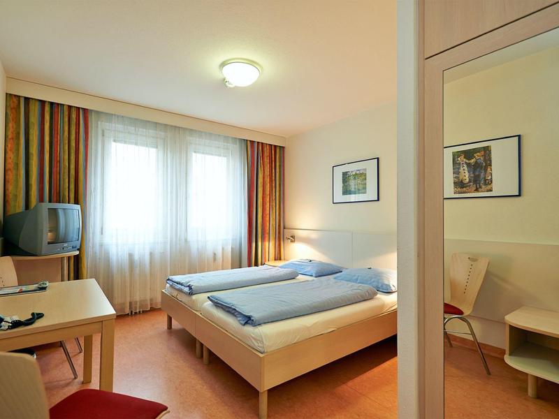 Lichte hotelkamer met tweepersoonsbed, bureau, tv en kledingkast met spiegel.